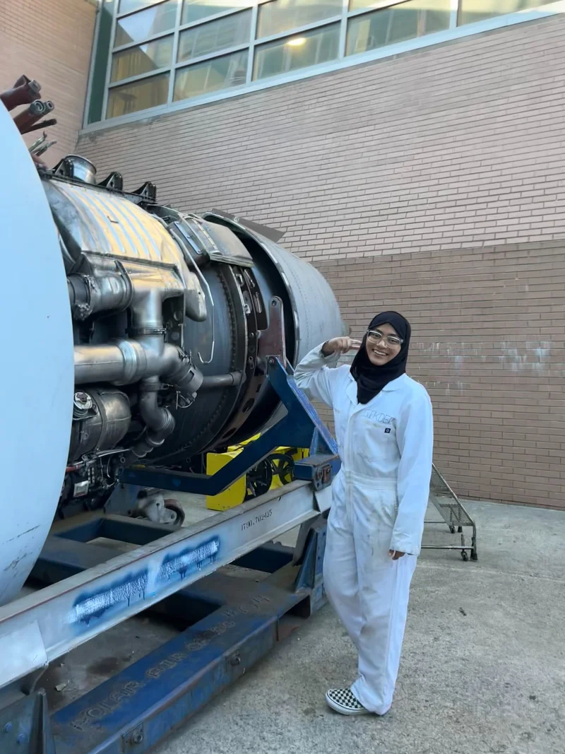 Zarin in coveralls standing beside an aircraft engine on a maintenance stand