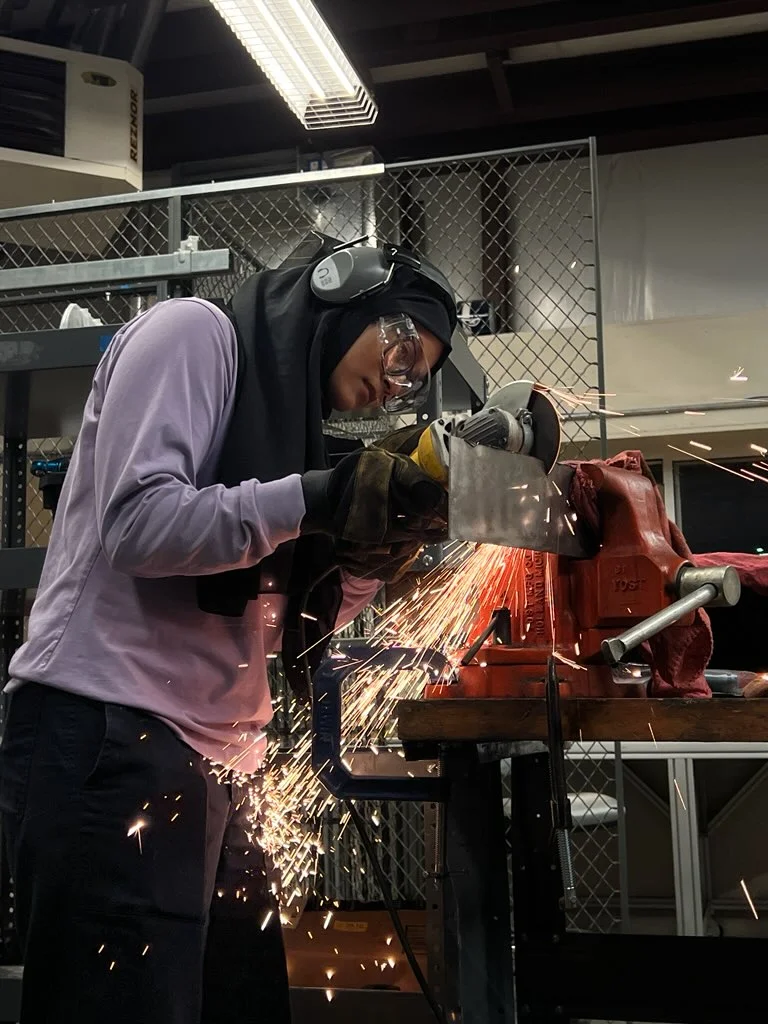 Zarin wearing eye and hearing protection while grinding metal at a workbench